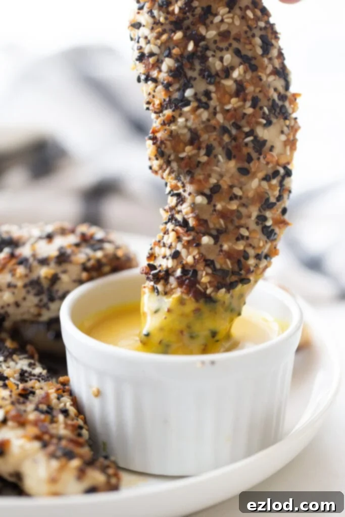 A close up of a chicken tender covered in everything bagel seasoning being dipped into maple mustard sauce.