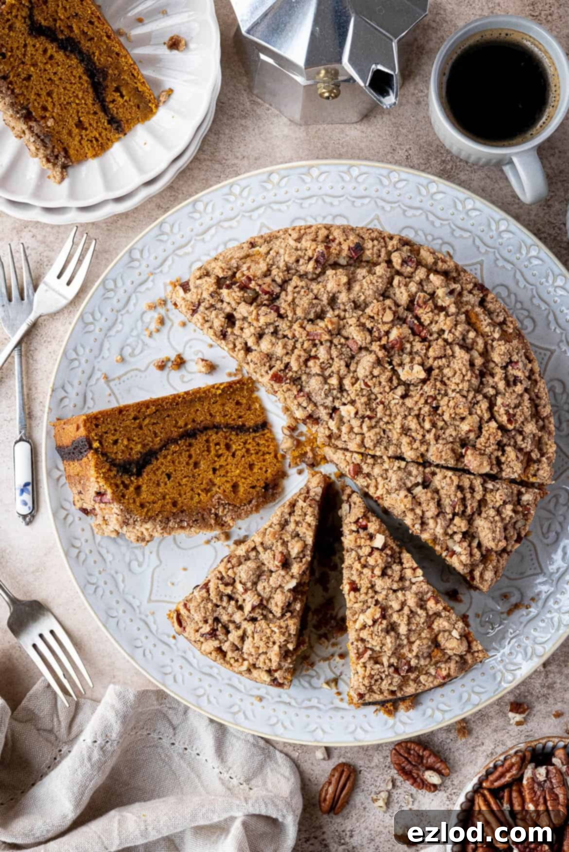 A beautifully sliced vegan pumpkin coffee cake on a white plate, with a coffee pot and a steaming cup of coffee in the background.