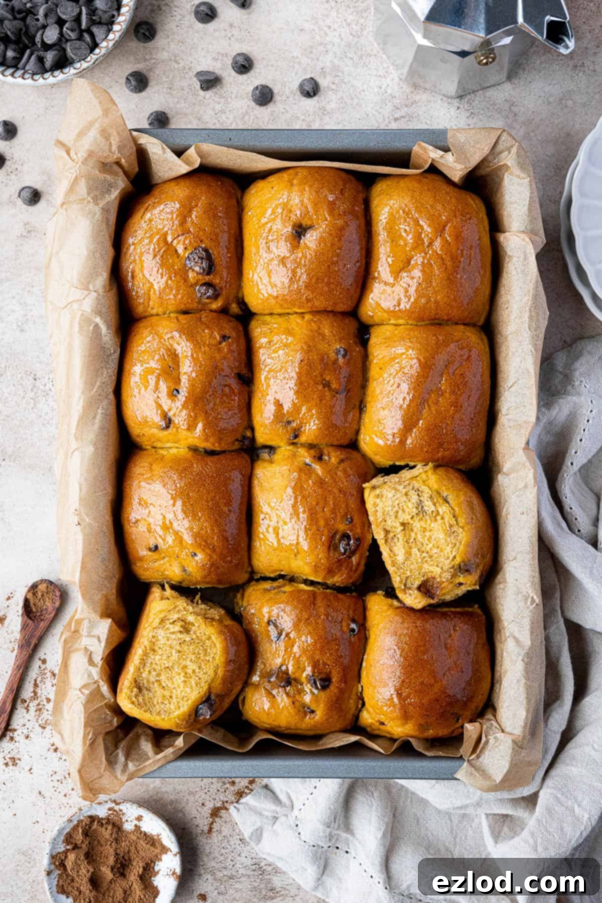 A tin filled with golden-brown chocolate chip pumpkin rolls, surrounded by bowls of chocolate chips and pumpkin spice, evoking a warm autumnal scene.