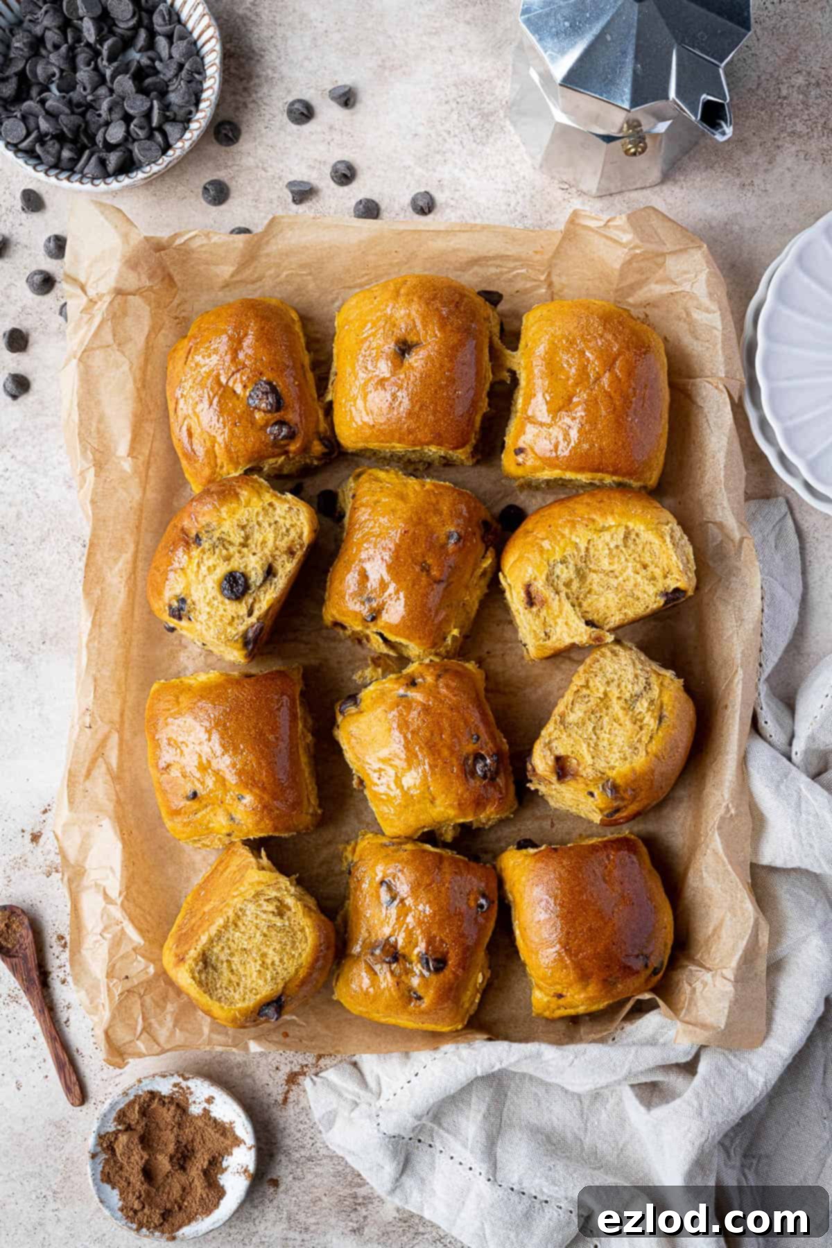A festive autumn scene featuring chocolate chip pumpkin rolls on baking paper, accompanied by a coffee pot, bowls of chocolate chips, and pumpkin spice.