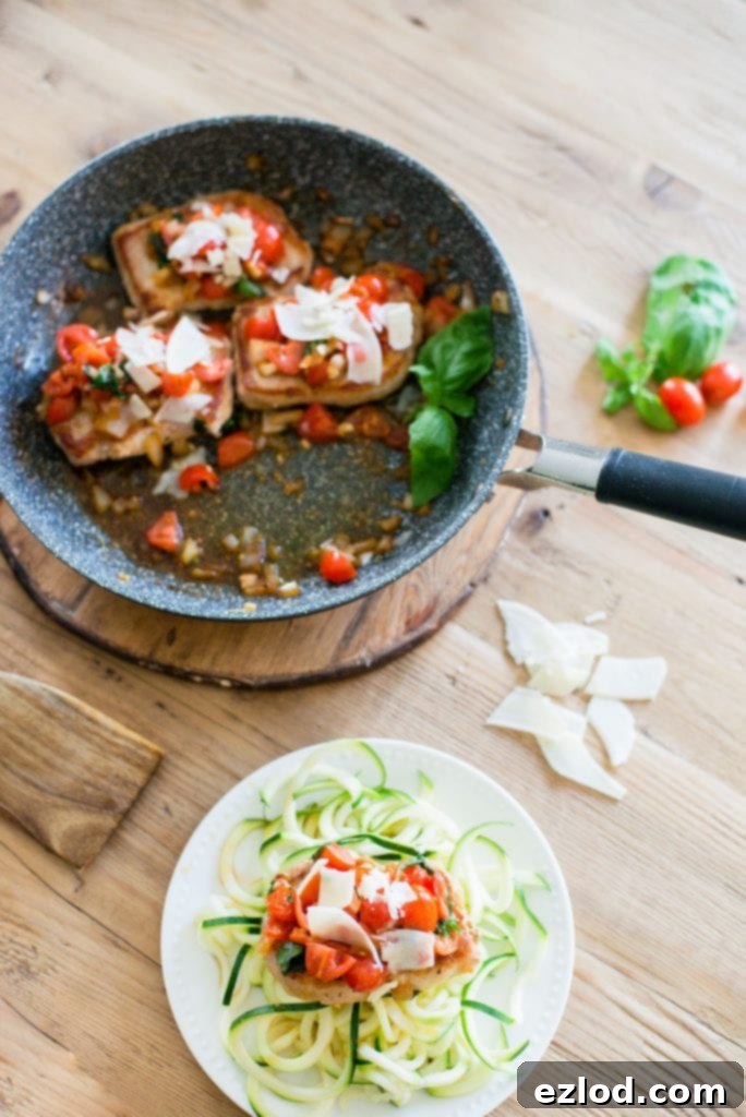 Skillet with three Italian Pork Chops topped with tomatoes, fresh basil, garlic, onions and parmesan cheese. A small white plate is nearby with a pork chop over zucchini noodles. 