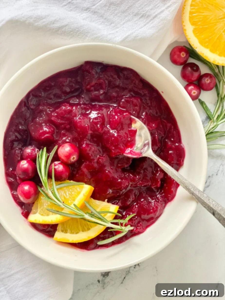 Overhead image: Homemade cranberry sauce in a white bowl, garnished with orange slices, whole cranberries, and sprigs of rosemary.
