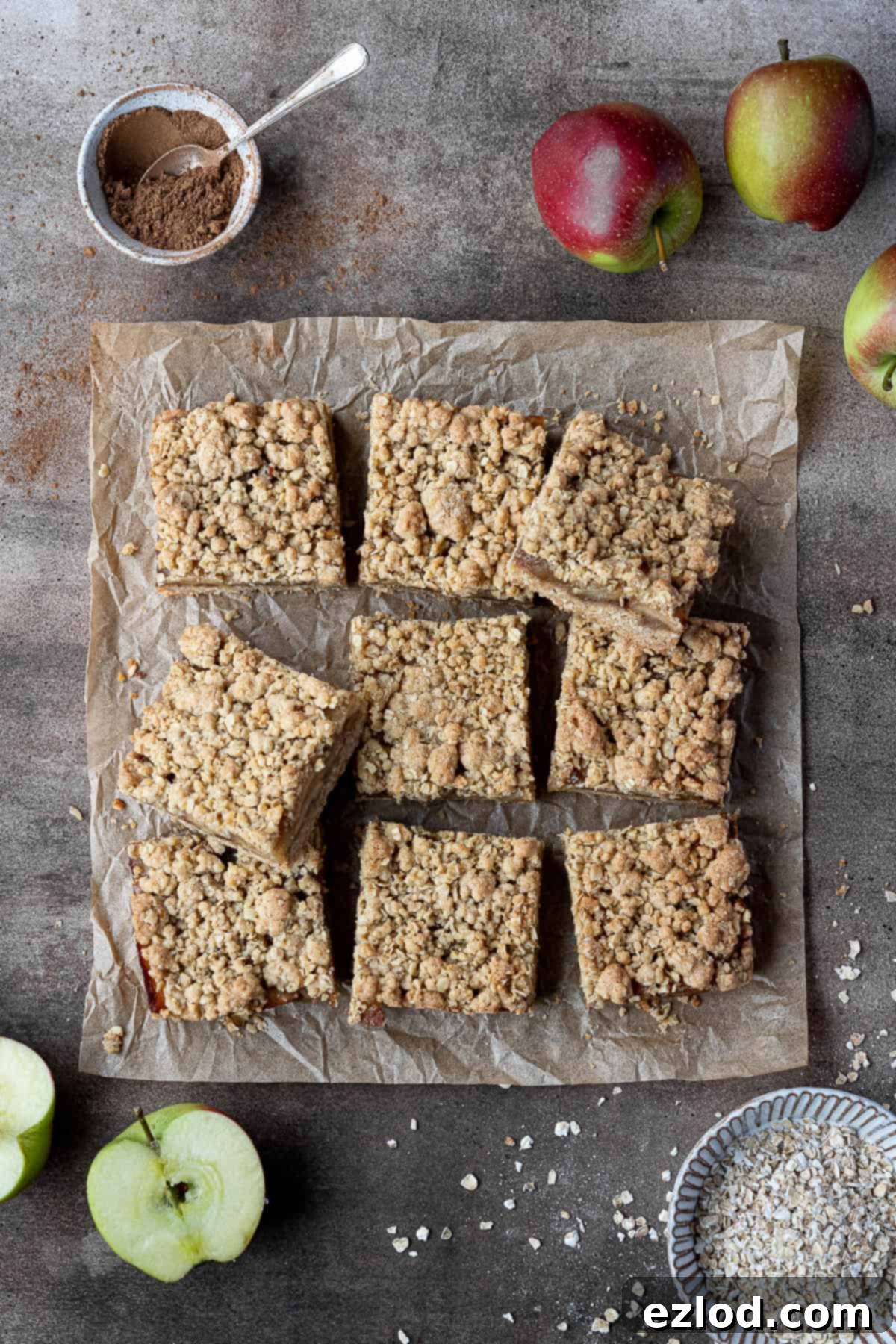 Vegan apple crumble bars on a sheet of baking parchment, surrounded by fresh apples, bowls of oats, and ground cinnamon, ready for baking.