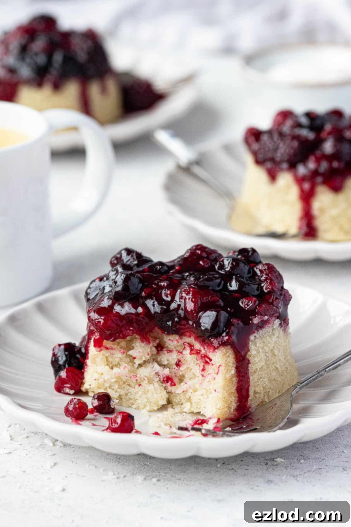 Three individual vegan berry and coconut steamed puddings on white plates, one with a fork revealing the layers of compote and sponge.