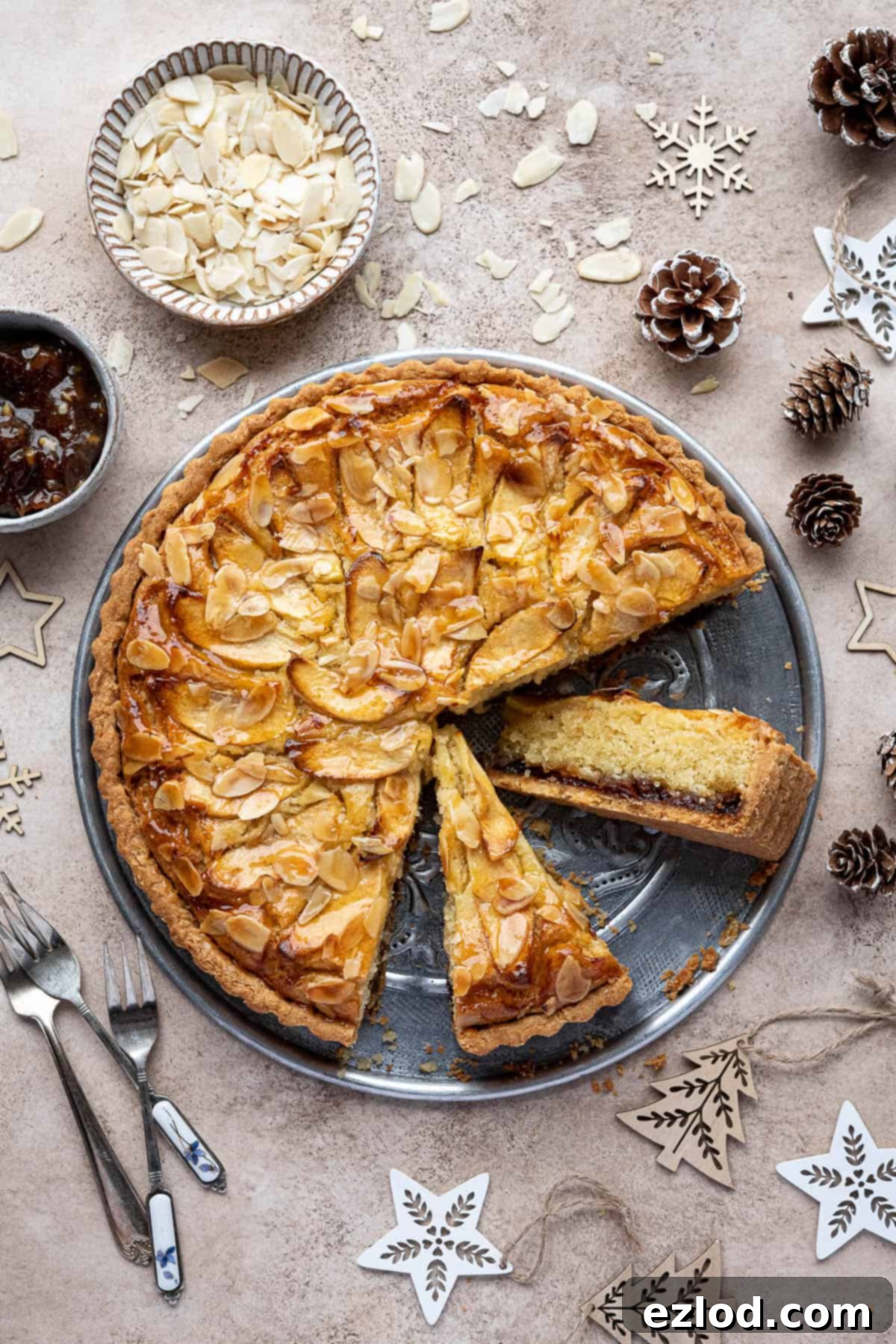 A beautifully baked vegan mincemeat frangipane tart with two slices already cut out, resting on a wooden surface surrounded by festive Christmas decorations, pinecones, and small bowls filled with flaked almonds and mincemeat.