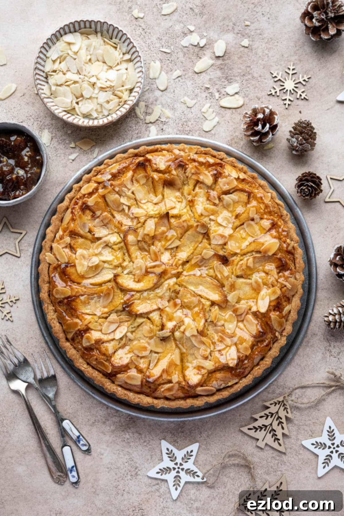 A festive scene featuring the vegan mincemeat frangipane tart on a metallic serving plate, surrounded by Christmas decorations, pinecones, and bowls of flaked almonds and mincemeat.