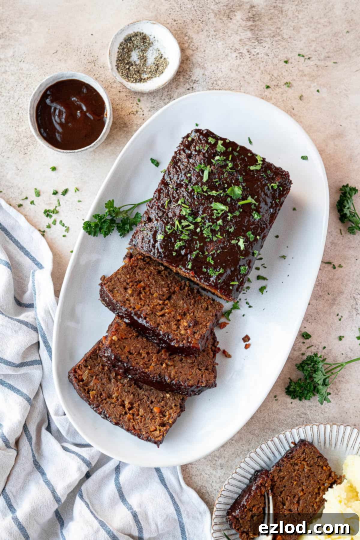 A perfectly sliced vegan seitan meatloaf on a plate, accompanied by bowls of rich barbecue sauce and freshly ground pepper.