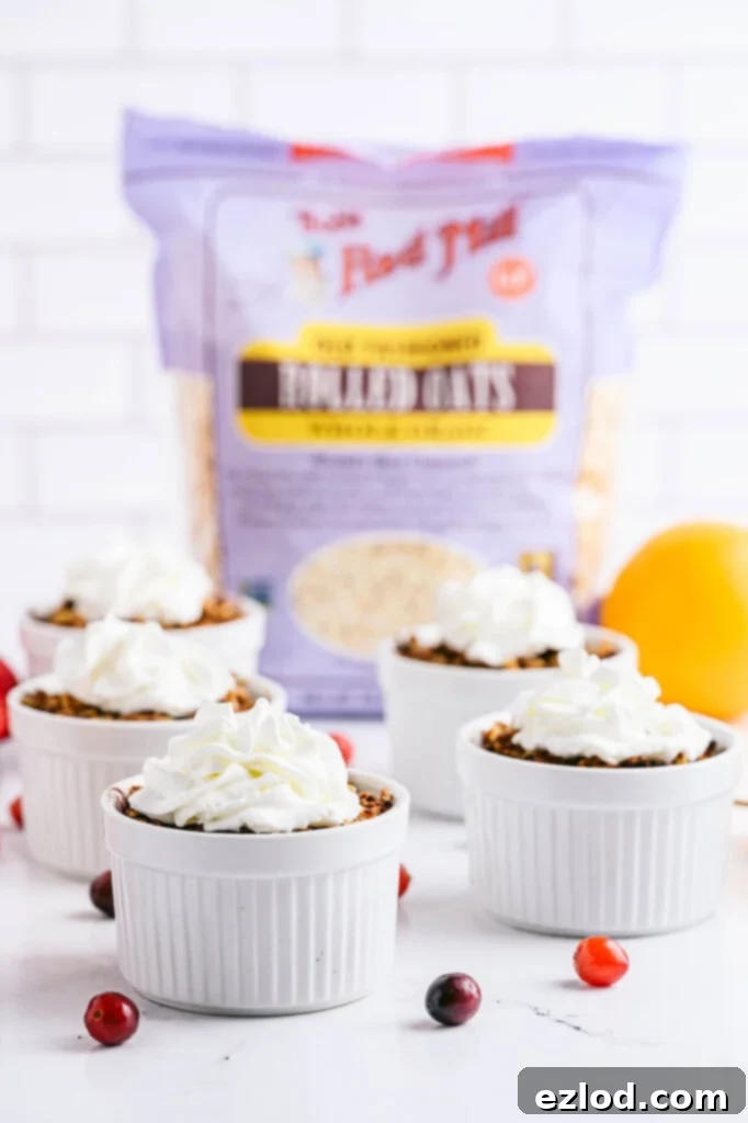Close up of four white ramekins with pear apple cranberry crisp topped with coconut whipped cream and with fresh cranberries and apples scattered around the table.  A bag of Bob's Red Mill Old Fashioned Oats sits in the background.