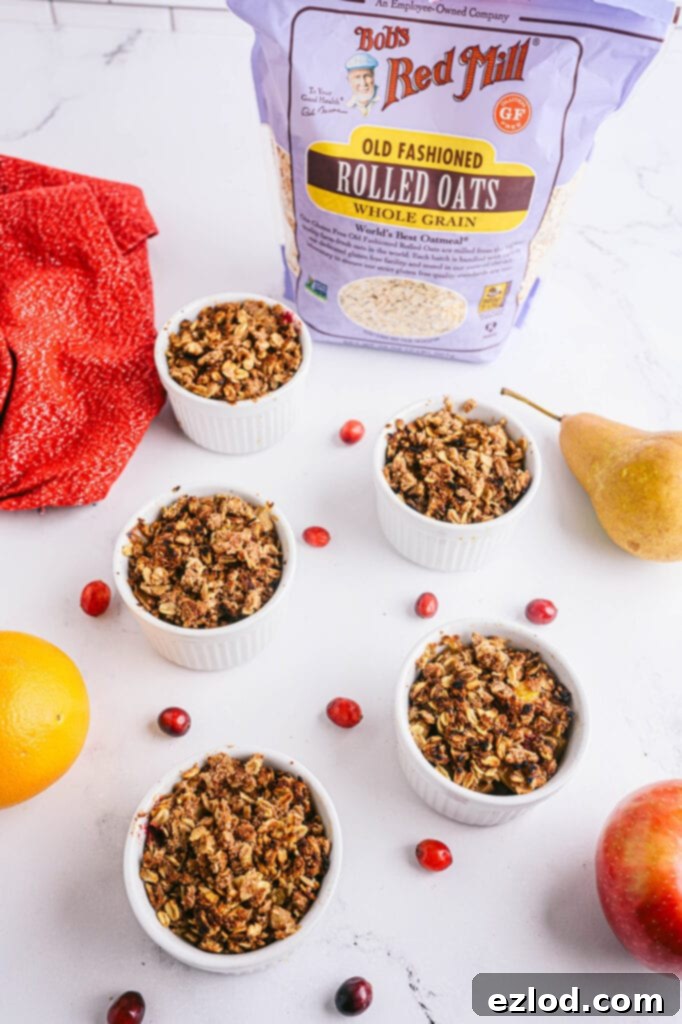Overhead shot of four white ramekins with pear apple cranberry crisp topped with coconut whipped cream and with fresh cranberries and apples scattered around the table. A bag of Bob's Red Mill Old Fashioned Oats sits in the background.