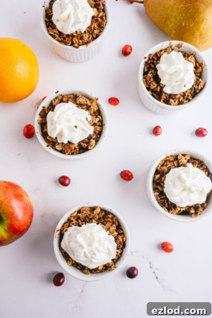 Overhead shot of four white ramekins with pear apple cranberry crisp topped with coconut whipped cream and with fresh cranberries and apples scattered around the table.