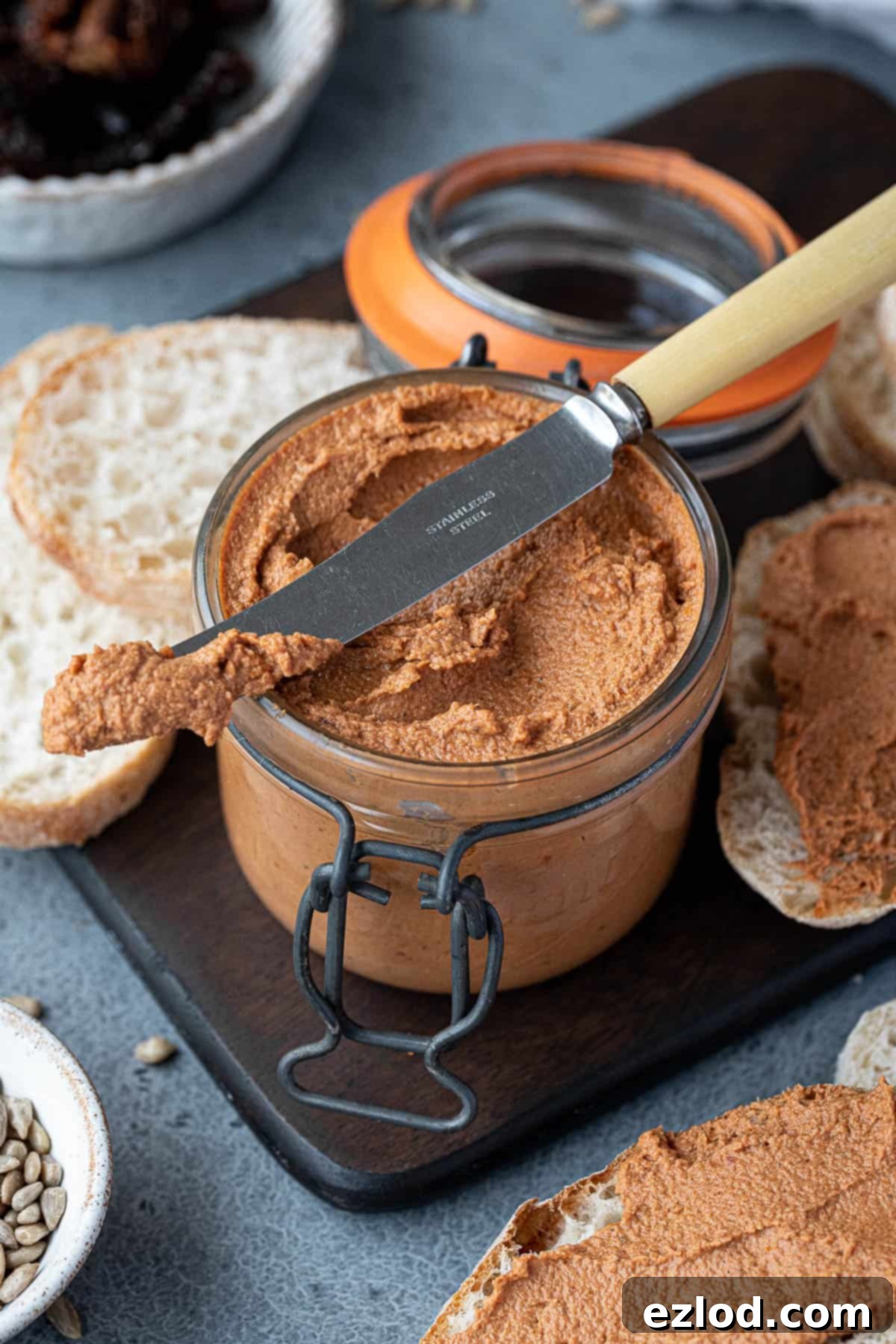 Vegan Tomato Sunflower Spread 3 Another view of a jar of tomato sunflower seed pâté on a wooden board, with a knife and sliced bread, highlighting its vibrant color and smooth texture.