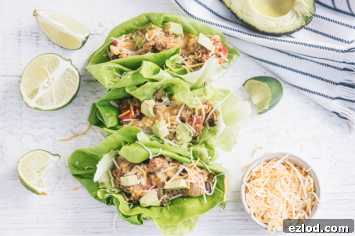 Three lettuce wraps filled with crock pot taco casserole on a white surface. Surrounding the wraps are a white and blue striped towel, half an avocado, sliced limes, and a ramekin full of shredded cheese.