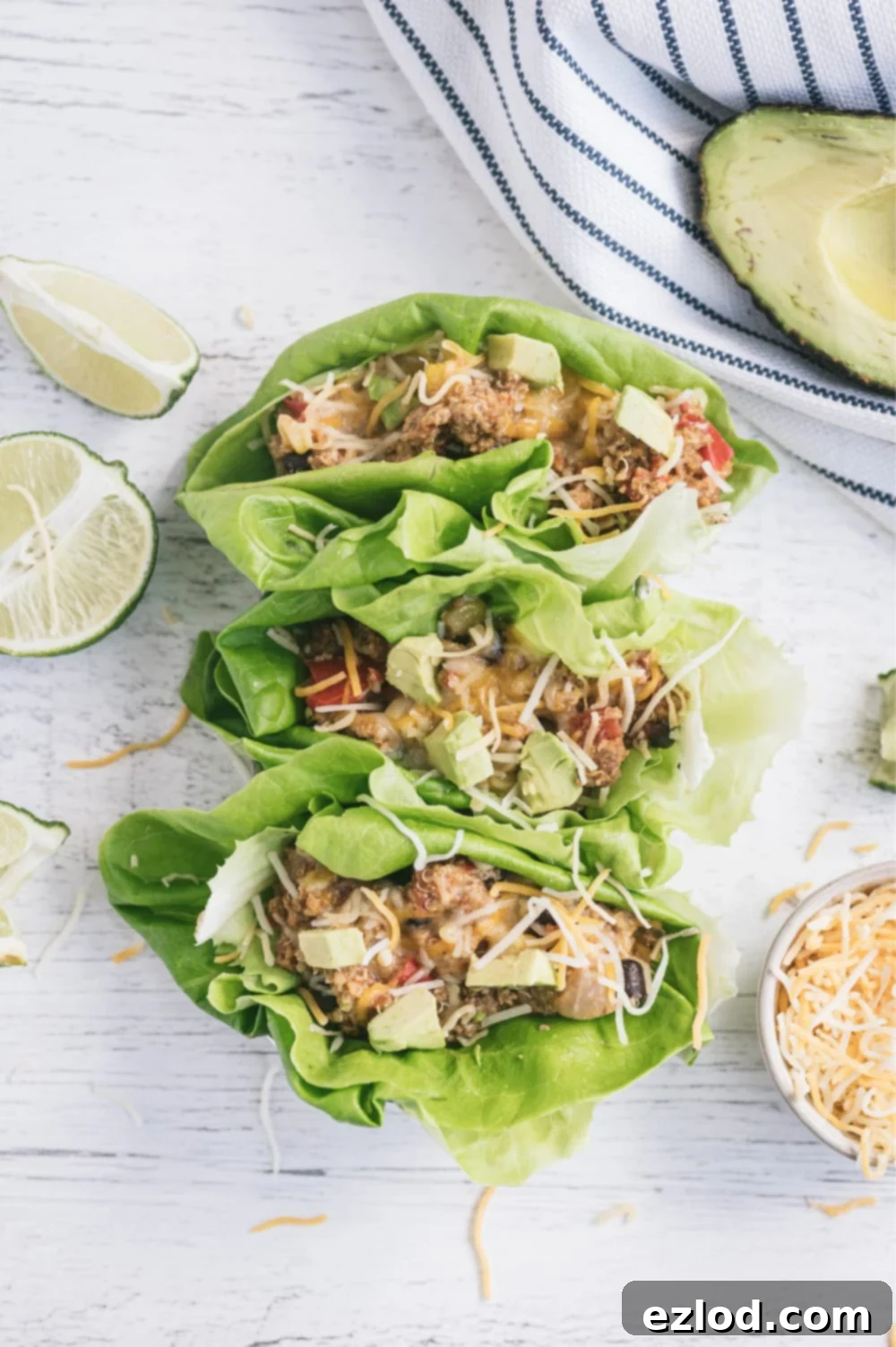 Overhead photo with lettuce wraps filled with Mexican crock pot taco casserole on a white surface. Surrounding the wraps are a white and blue striped towel, half an avocado, sliced limes, and a ramekin full of shredded cheese.