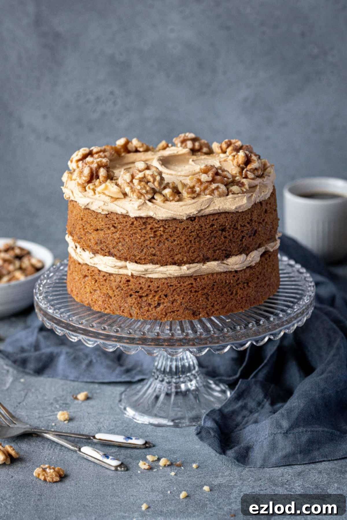 Gluten-free vegan coffee and walnut cake on a glass cake stand with a bowl of walnuts and cups of coffee.