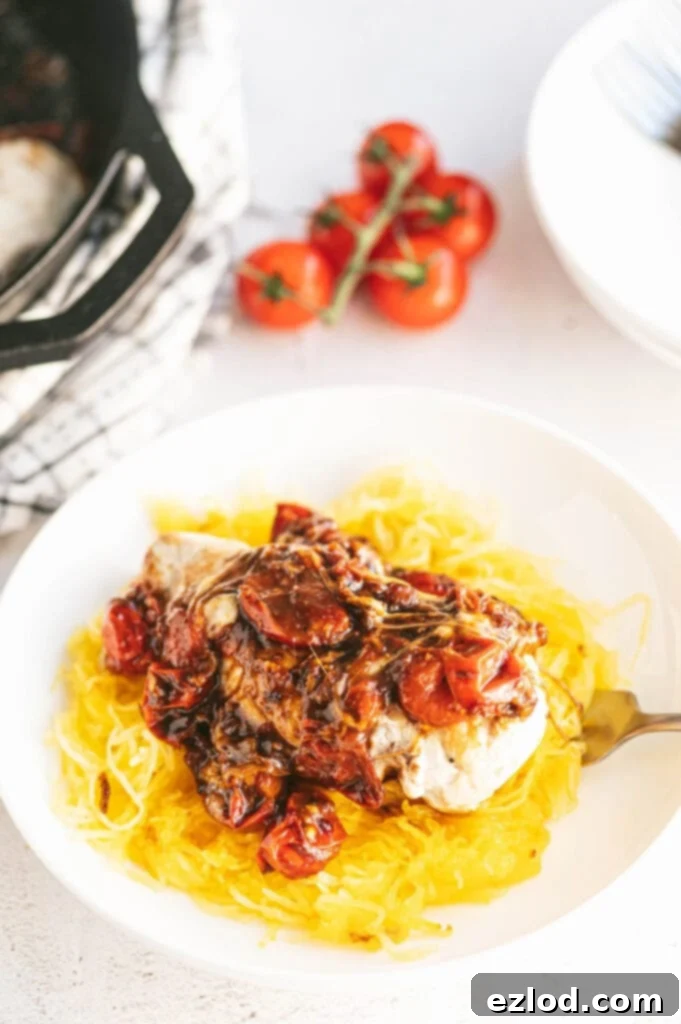 One skillet Caprese Chicken made with balsamic vinegar, juicy tomatoes, onions, and topped with mozzarella cheese. The chicken and tomatoes are over a generous serving of spaghetti squash in a white bowl. In the background, out of focus, are vine ripe tomatoes, plates, and the skillet with more chicken to serve.