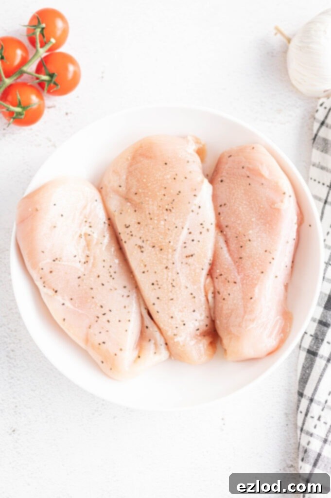 Overhead photo of three seasoned raw chicken breasts in a white bowl with whole tomatoes and garlic on the side.