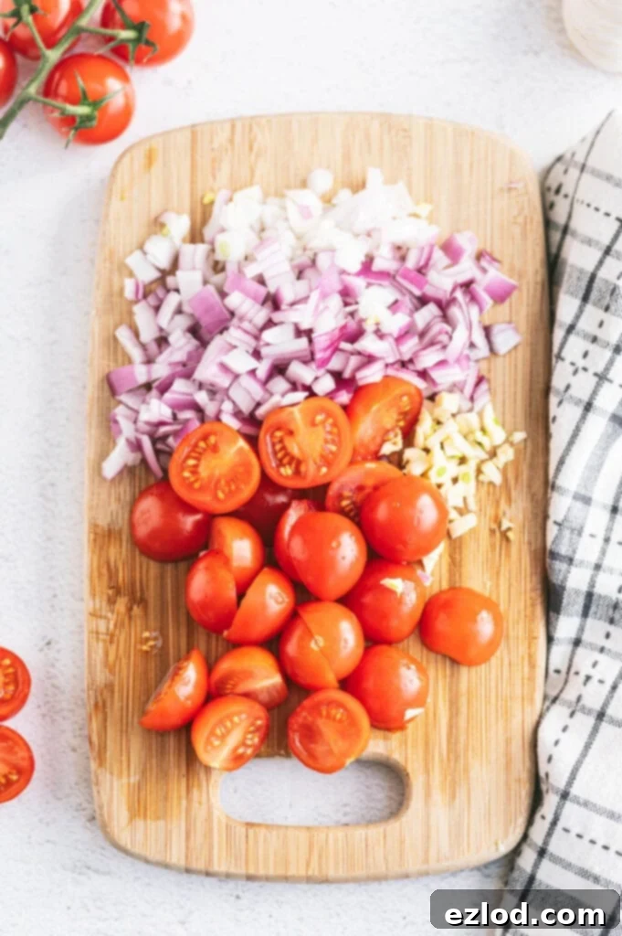 Overhead photo of halved cherry tomatoes, diced red onion, and minced garlic on a cutting board ready to make a caprese chicken recipe.