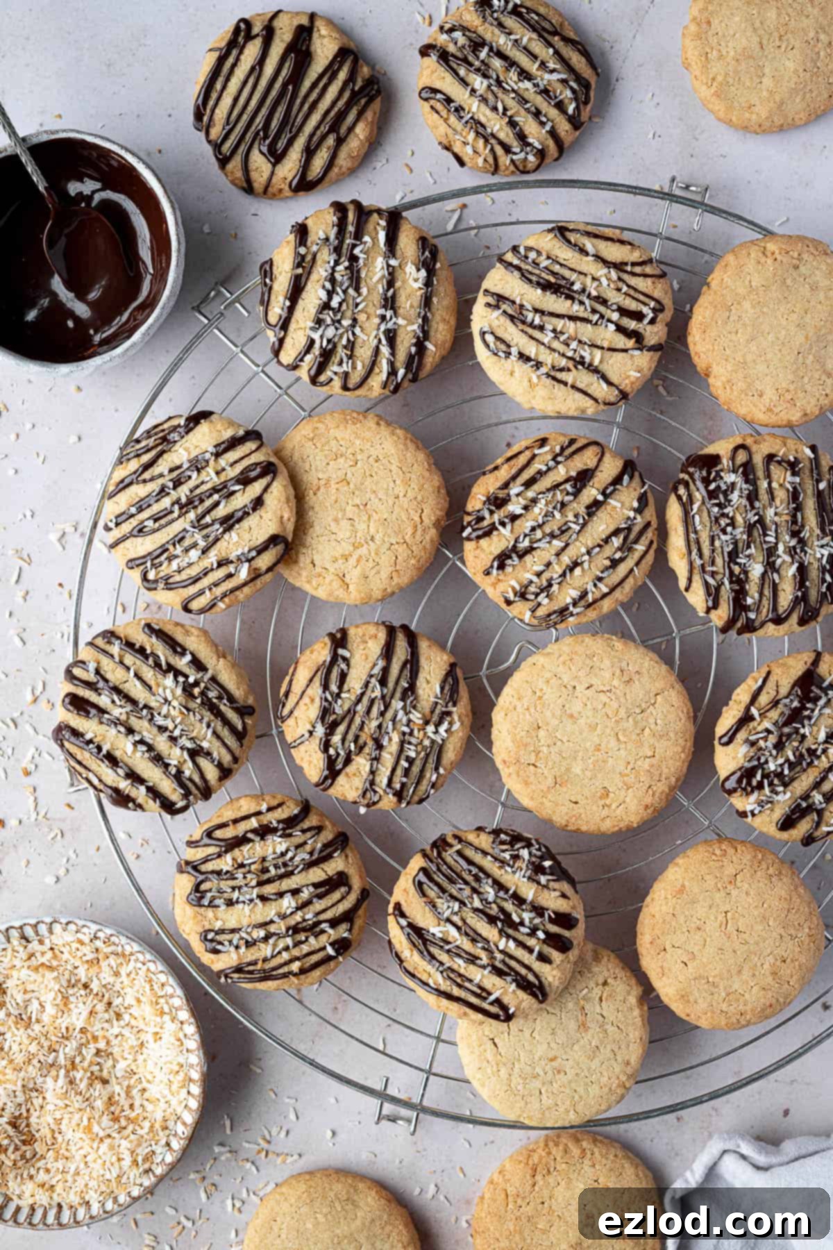 Vegan coconut cookies on a wire cooling rack with bowls of melted chocolate and toasted coconut.