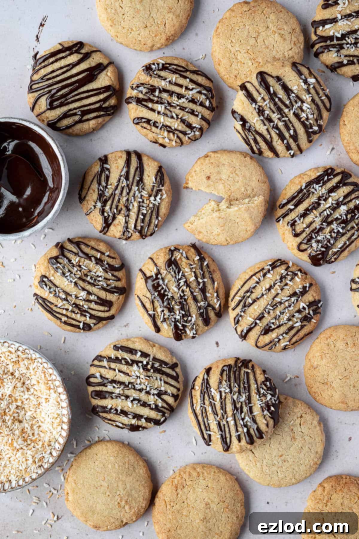 Vegan coconut cookies on a marble surface with bowls of melted chocolate and toasted coconut.