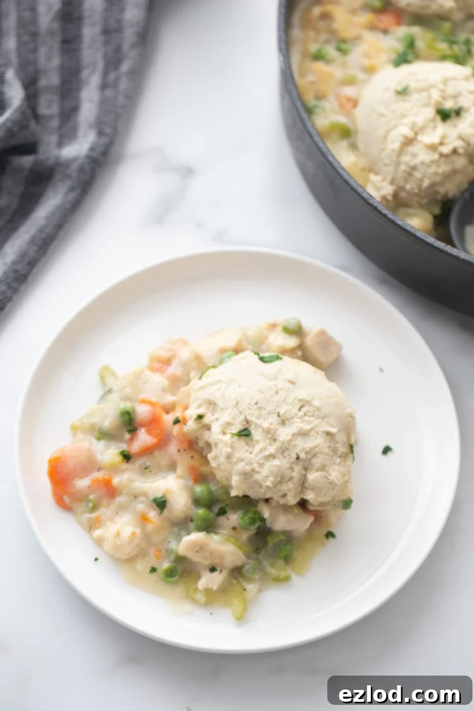 Skillet chicken pot pie with gluten-free biscuit topping piled on a white plate on a white and gray marble table. The skillet is in the upper right hand corner of the shot. 
