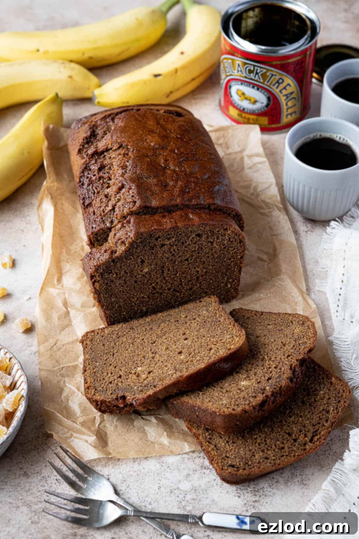Banana gingerbread on a sheet of baking paper with cups  of coffee, a tin of treacle, bananas, forks and crystallised ginger.