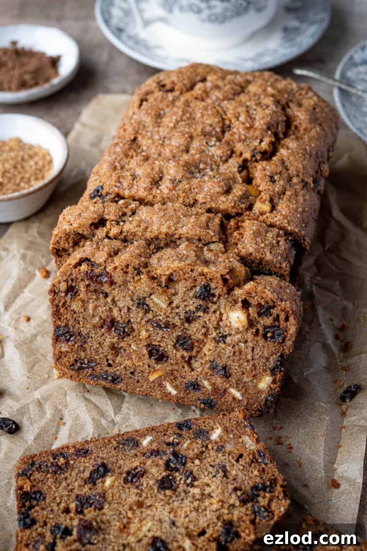 Sliced vegan fruit cake loaf arranged with bowls of sugar and spice, and a teacup in the background, inviting a tea time treat.