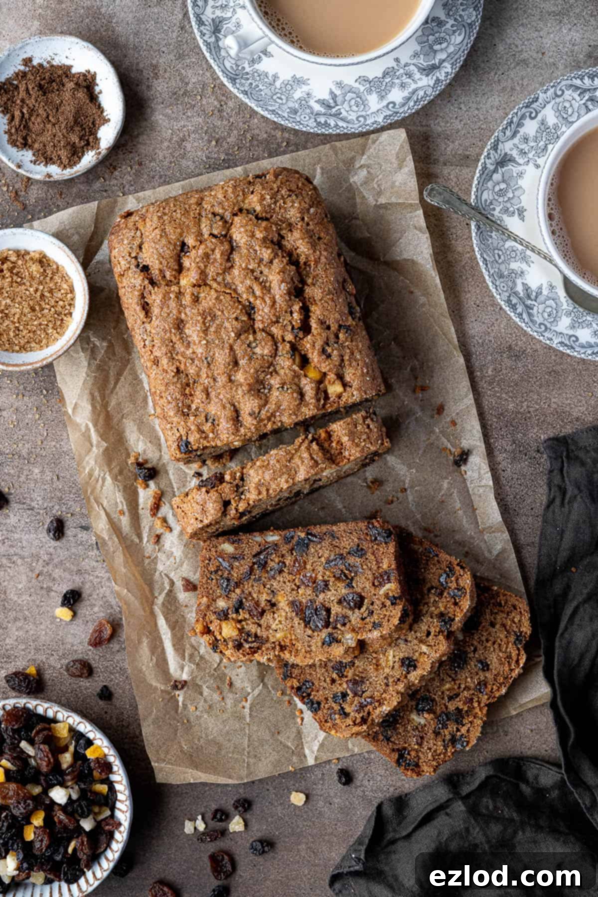Ingredients for vegan fruit cake loaf laid out on a brown surface, including dried fruit, sugar, spices, and a vegan butter block, with cups of tea in the background.