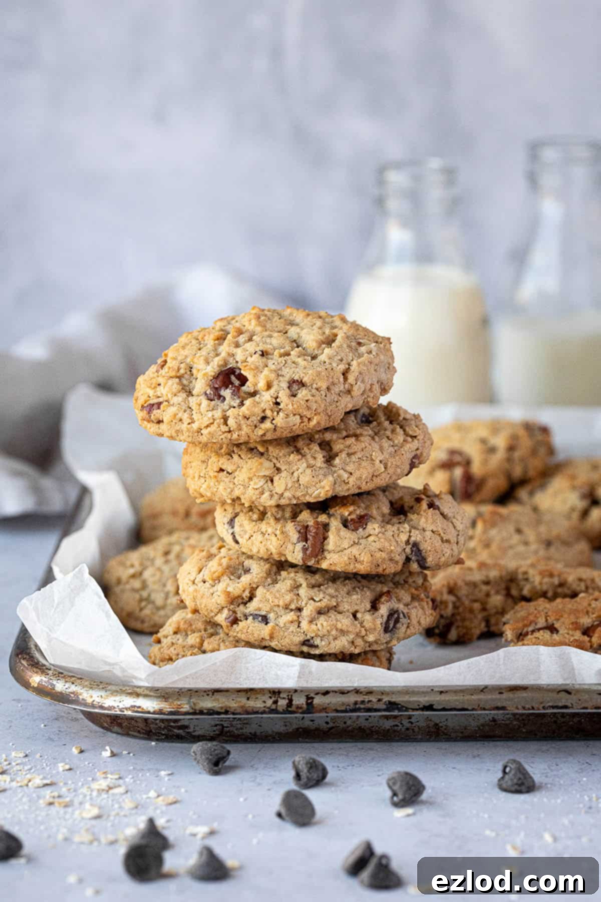 A stack of golden brown vegan peanut butter oatmeal cookies, studded with chocolate chips and pecans, presented on a rustic wooden tray with bottles of plant-based milk and scattered chocolate chips in the background.
