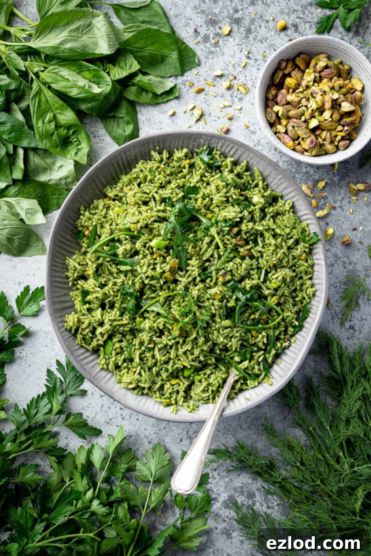A vibrant green herby rice salad with pistachios in a grey bowl, garnished with fresh herbs and surrounded by a bowl of pistachios.