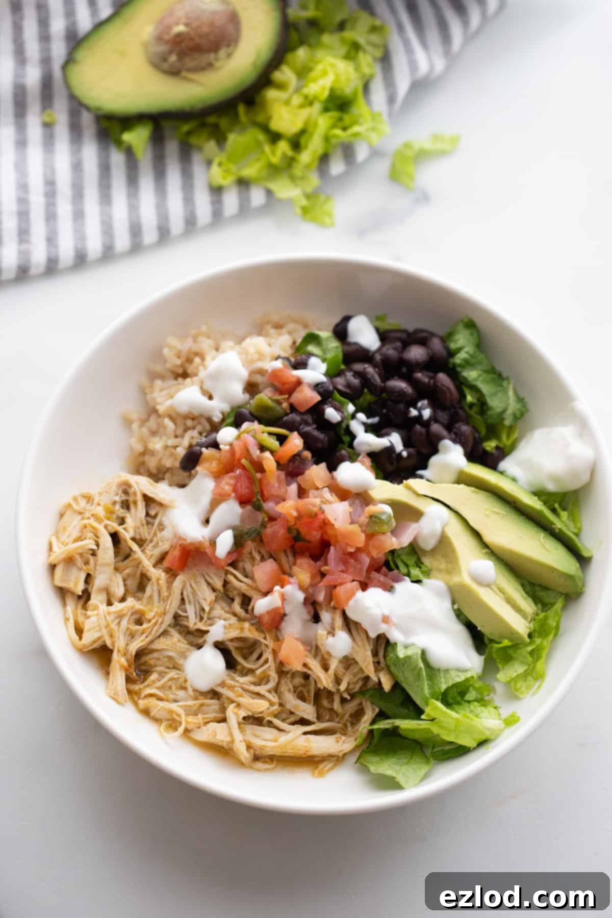 Effortless Salsa Verde Chicken Instant Pot or Slow Cooker 2 A vibrant bowl of shredded salsa verde chicken, generously topped with creamy Greek yogurt, fluffy brown rice, fresh pico de gallo, crisp greens, and sliced avocado, all presented on a pristine white table alongside a charming blue striped napkin.