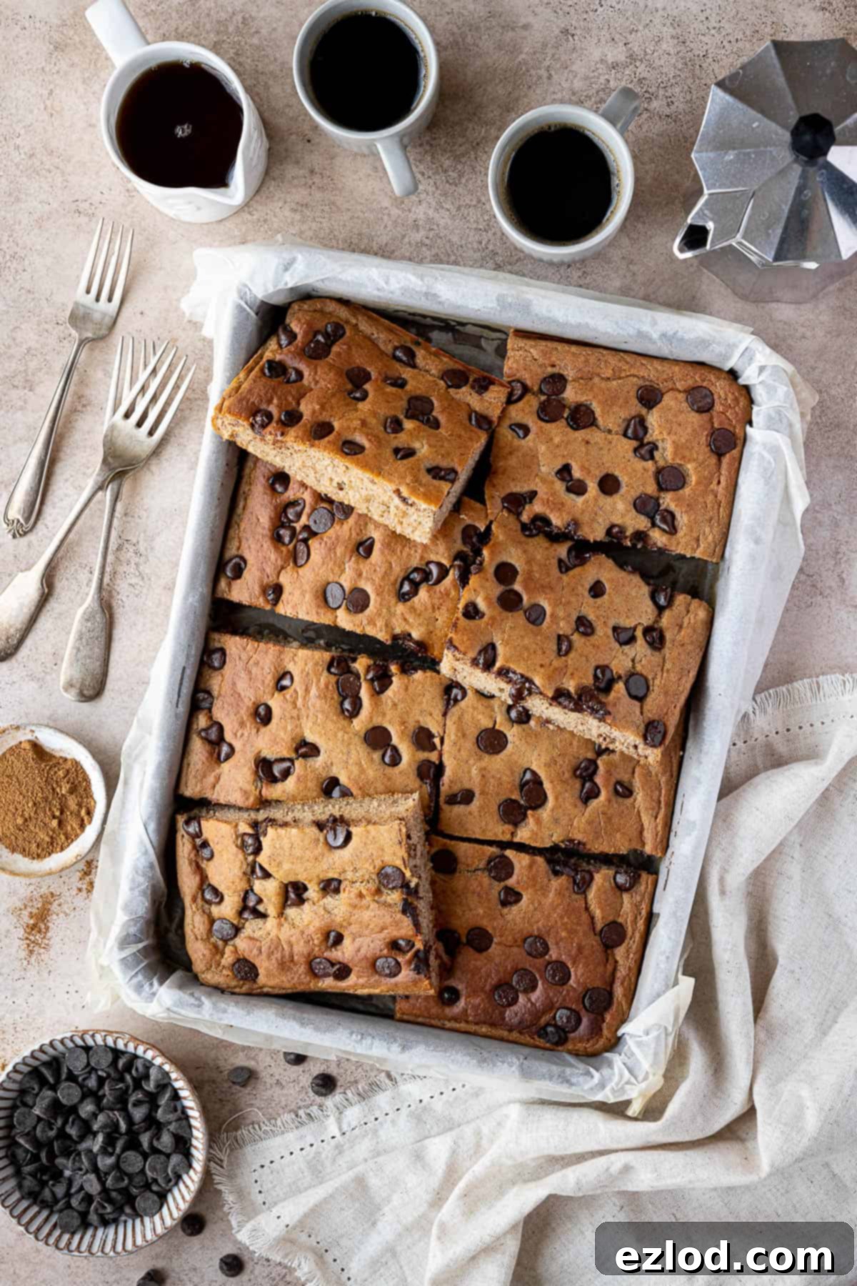 Sheet pan banana pancakes sliced into squares in a tin with cups of coffee, a jug of maple syrup and a bowl of chocolate chips.