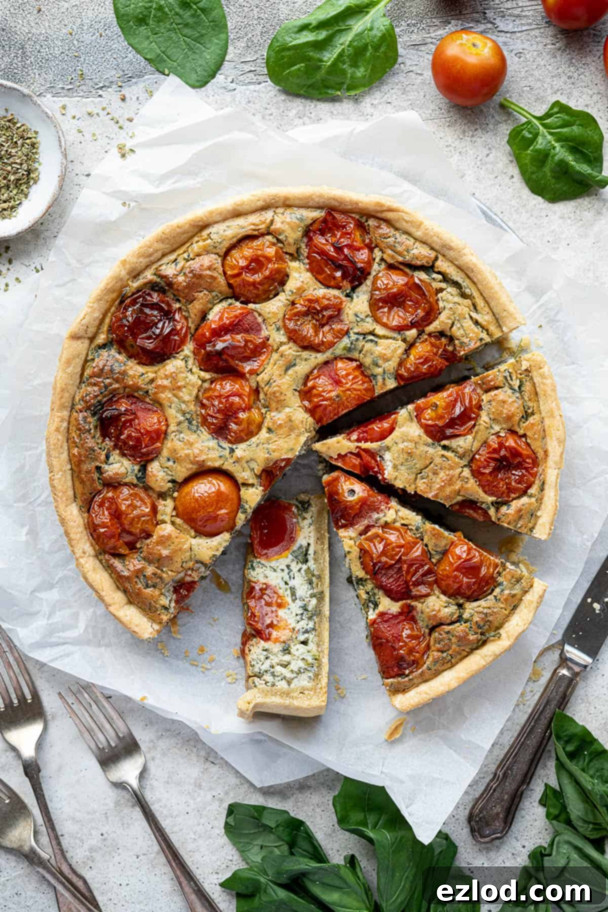 A beautifully sliced vegan tomato spinach quiche on baking parchment, surrounded by fresh basil leaves, vibrant spinach, and ripe cherry tomatoes.