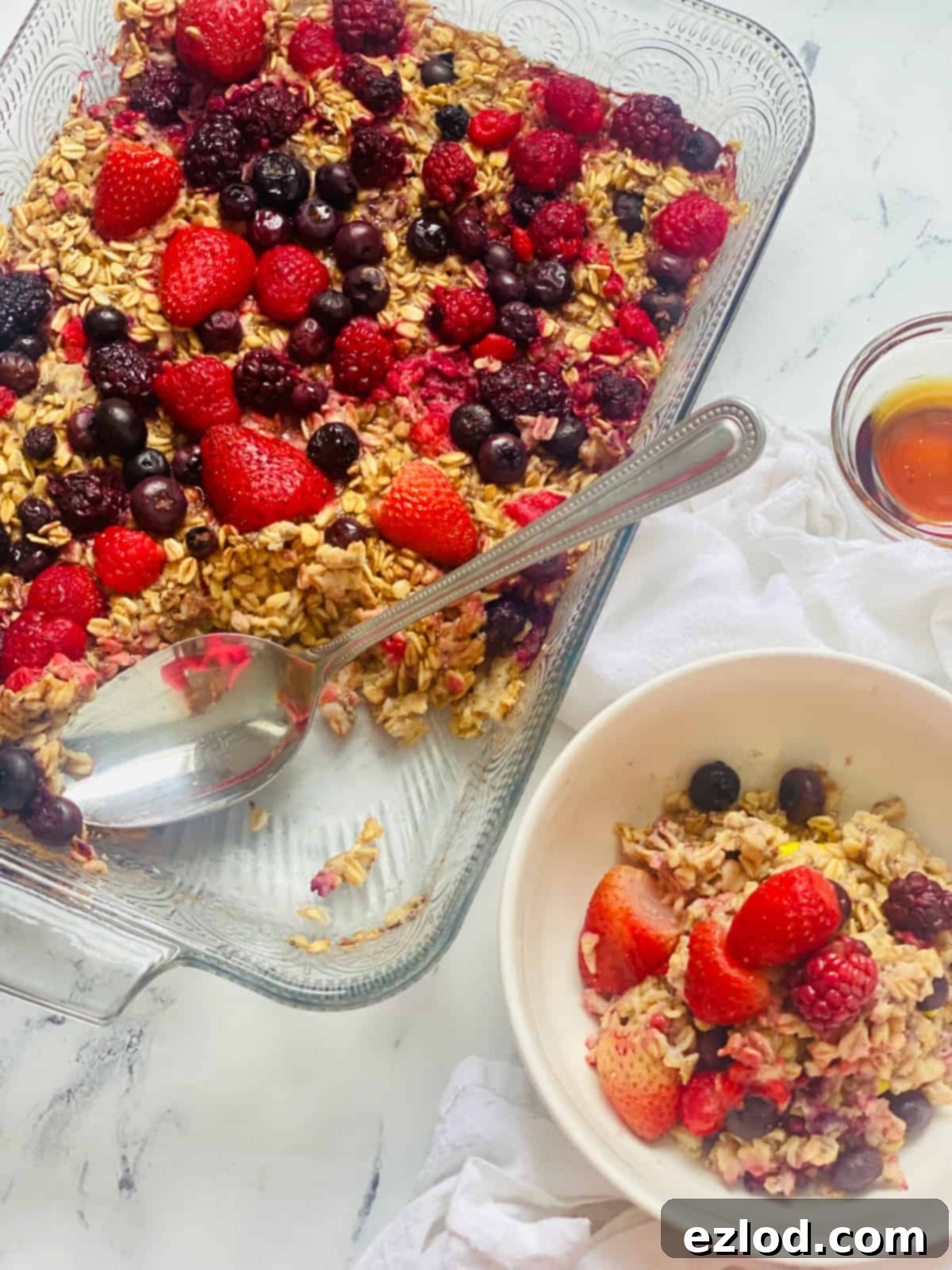 A close-up shot of a one-pan baked oatmeal dish with vibrant berries, next to a neatly portioned bowl on a marble countertop, highlighting its aesthetic appeal and ease of serving. 