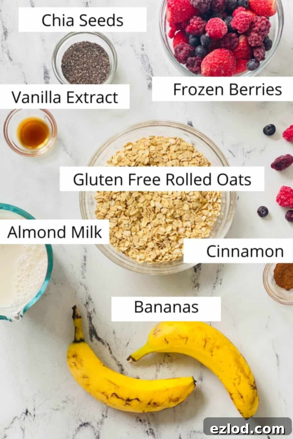 A flat lay arrangement of fresh ingredients for One Pan Baked Oatmeal laid out on a clean table. Each item is clearly labeled with text: ripe bananas, gluten-free oats, pure vanilla extract, a mix of frozen berries, unsweetened almond milk, and fragrant cinnamon, showcasing the simplicity of the recipe. 