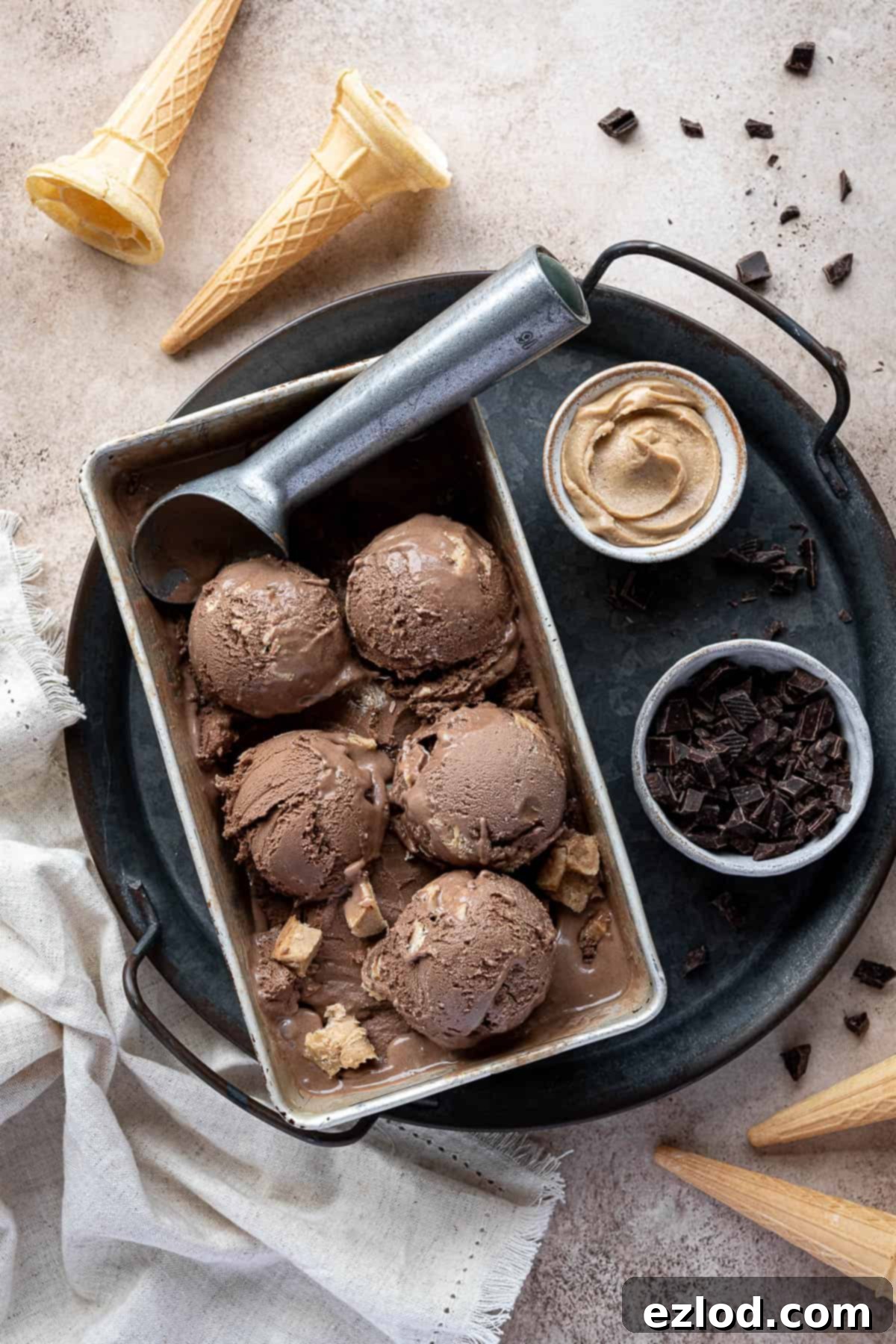 Scoops of vegan chocolate peanut butter ice cream in a loaf tin on a metal tray with ice cream cones annd bowls of chocolate and peanut butter.