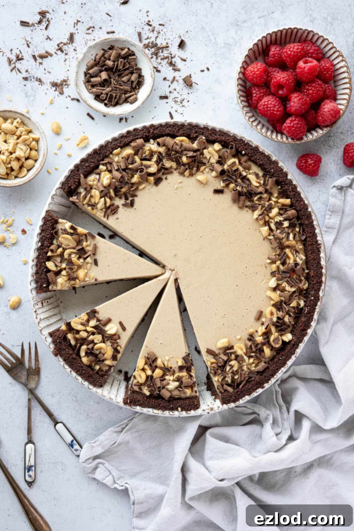 The whole vegan peanut butter pie, beautifully sliced, is displayed amidst small bowls of fresh raspberries, roasted peanuts, and chocolate, creating a vibrant and appealing scene for dessert enthusiasts, ready for social media sharing.