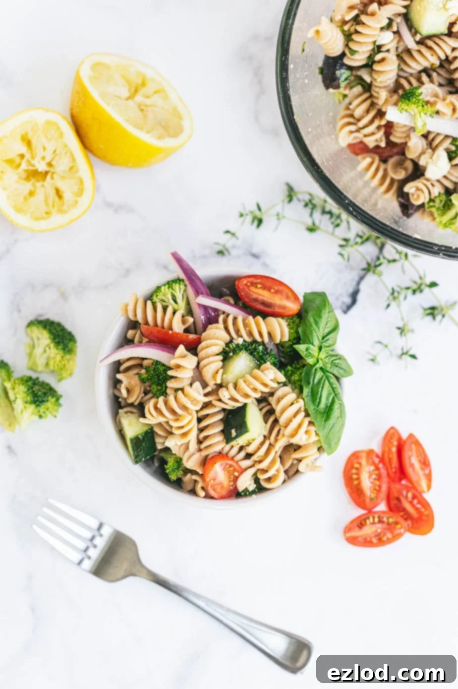 Overhead photo of an individual bowl of Greek pasta salad with a large glass serving bowl in the corner of the frame. Halved grape tomatoes, chopped broccoli, lemon halves, fresh thyme, and a fork are placed around the bowls.