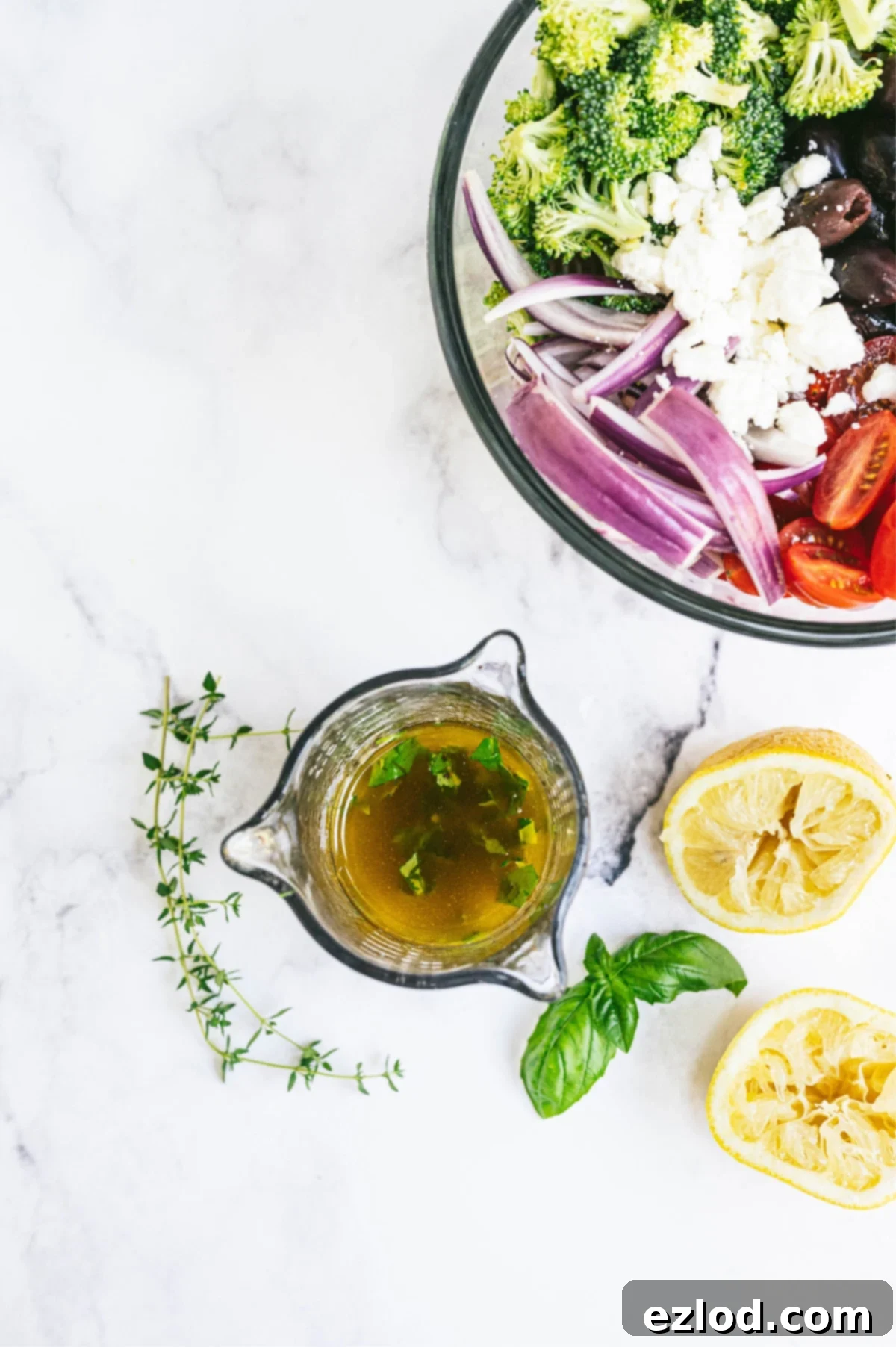 Overhead photo of glass container full of dressing, squeezed lemon halves, and a large glass bowl full of veggies and feta cheese.