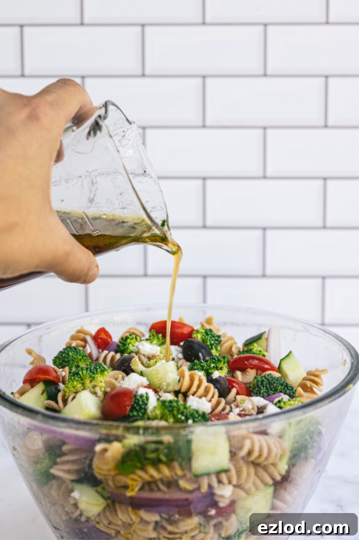 A hand pouring dressing from a glass container into a bowl bowl of Greek pasta salad with white subway tiles in the background.