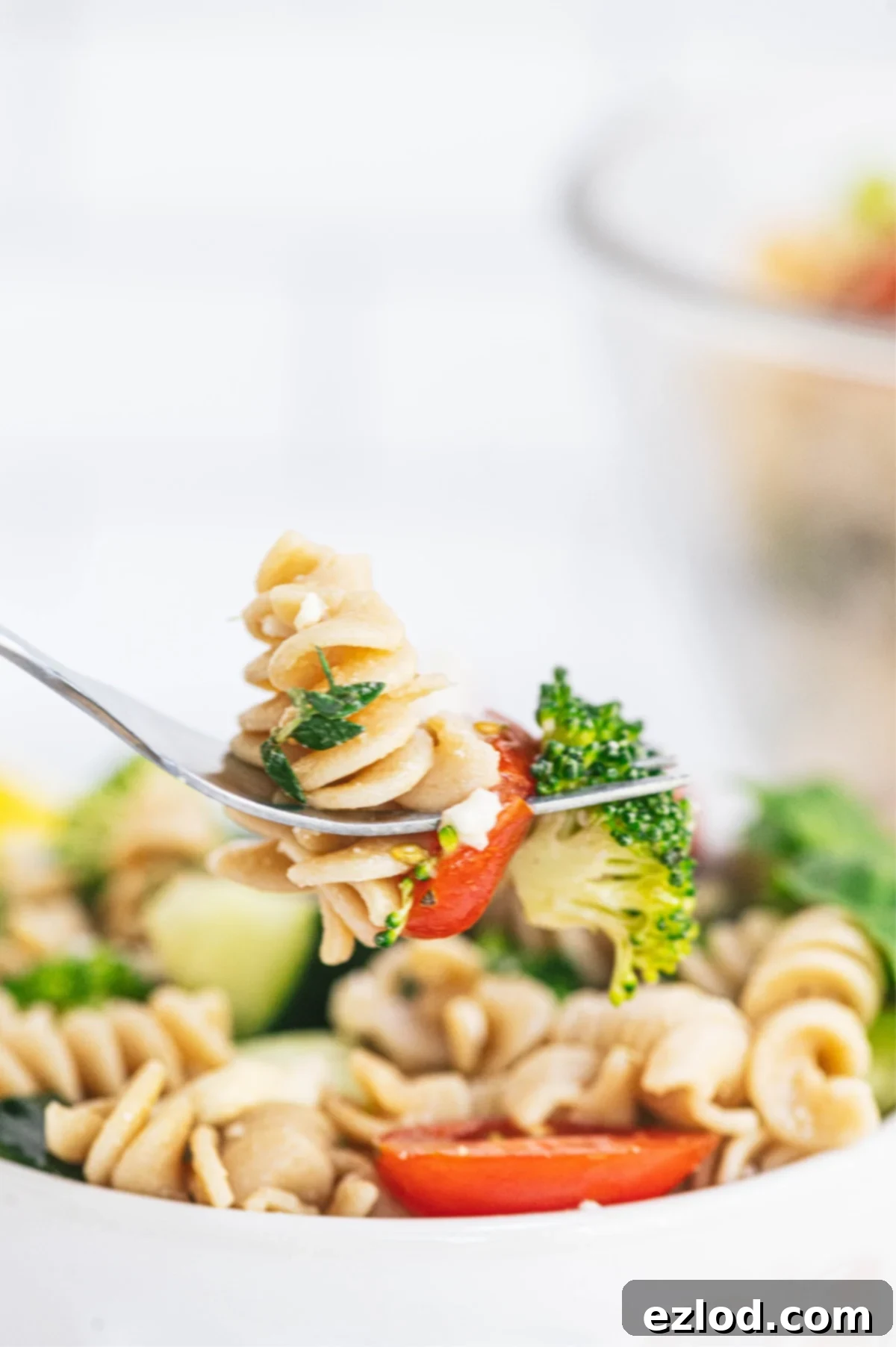 A fork full of pasta, broccoli, tomatoes and feta with a bowl of the best pasta salad in the background, slightly out of focus.
