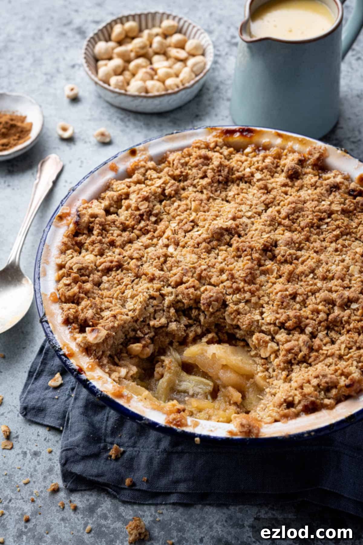 Close up of a dish of rhubarb apple crumble with a jug of custard and bowls of hazelnuts and cinnamon.