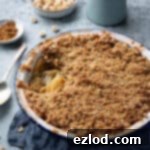 Rhubarb apple crumble in a round baking dish with a jug of custard and bowlds of hazelnuts and cinnamon.