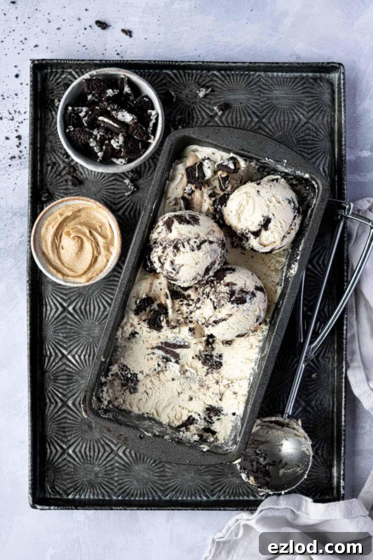 No-churn peanut butter Oreo ice cream in a loaf tin on a tray with an ice cream scoop and bowls of peanut butter and crushed Oreos.