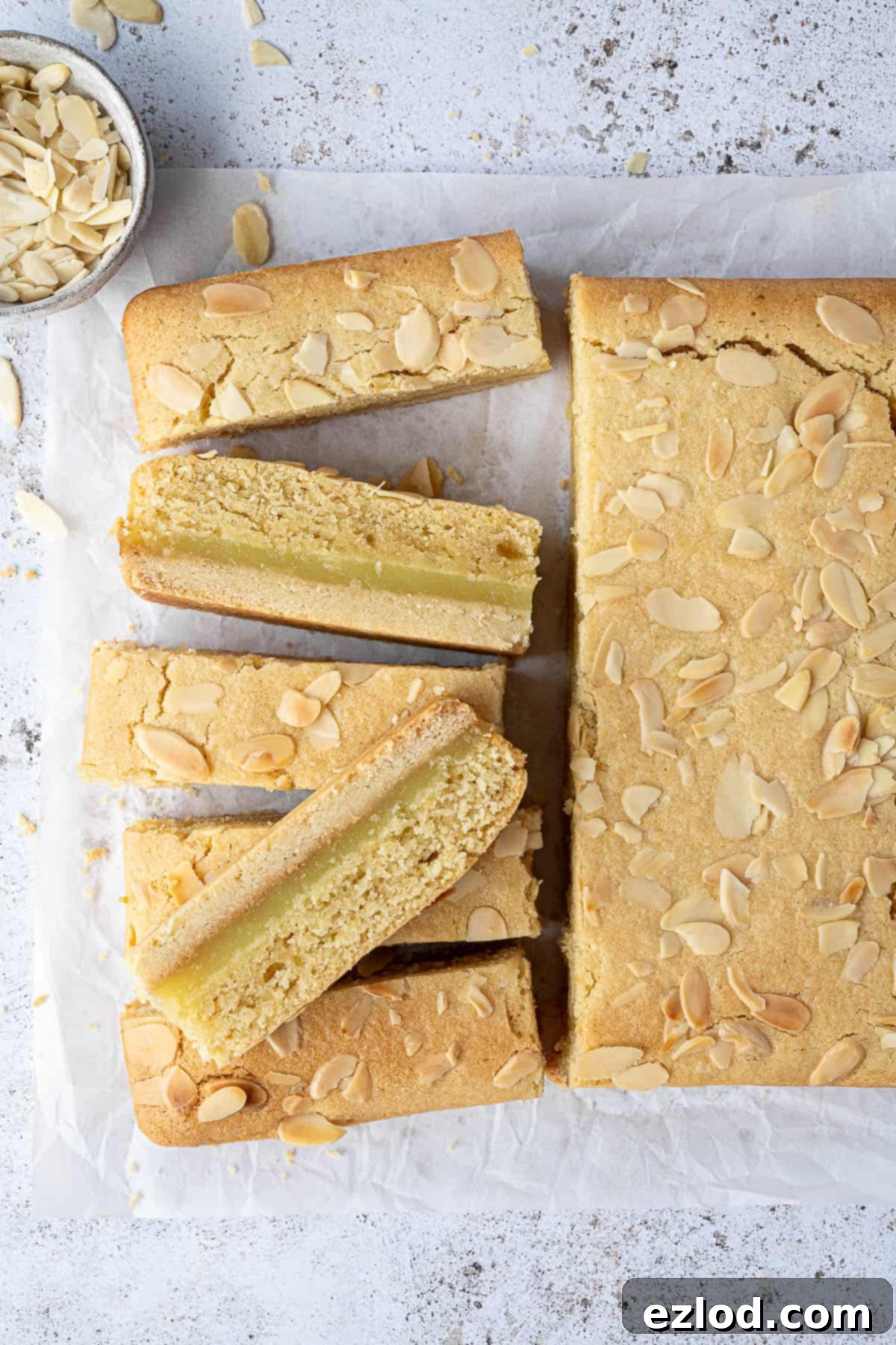 Zesty Vegan Bakewell Bliss 10 Vegan lemon Bakewell slices arranged on baking parchment with a small bowl of flaked almonds, ready to be enjoyed.