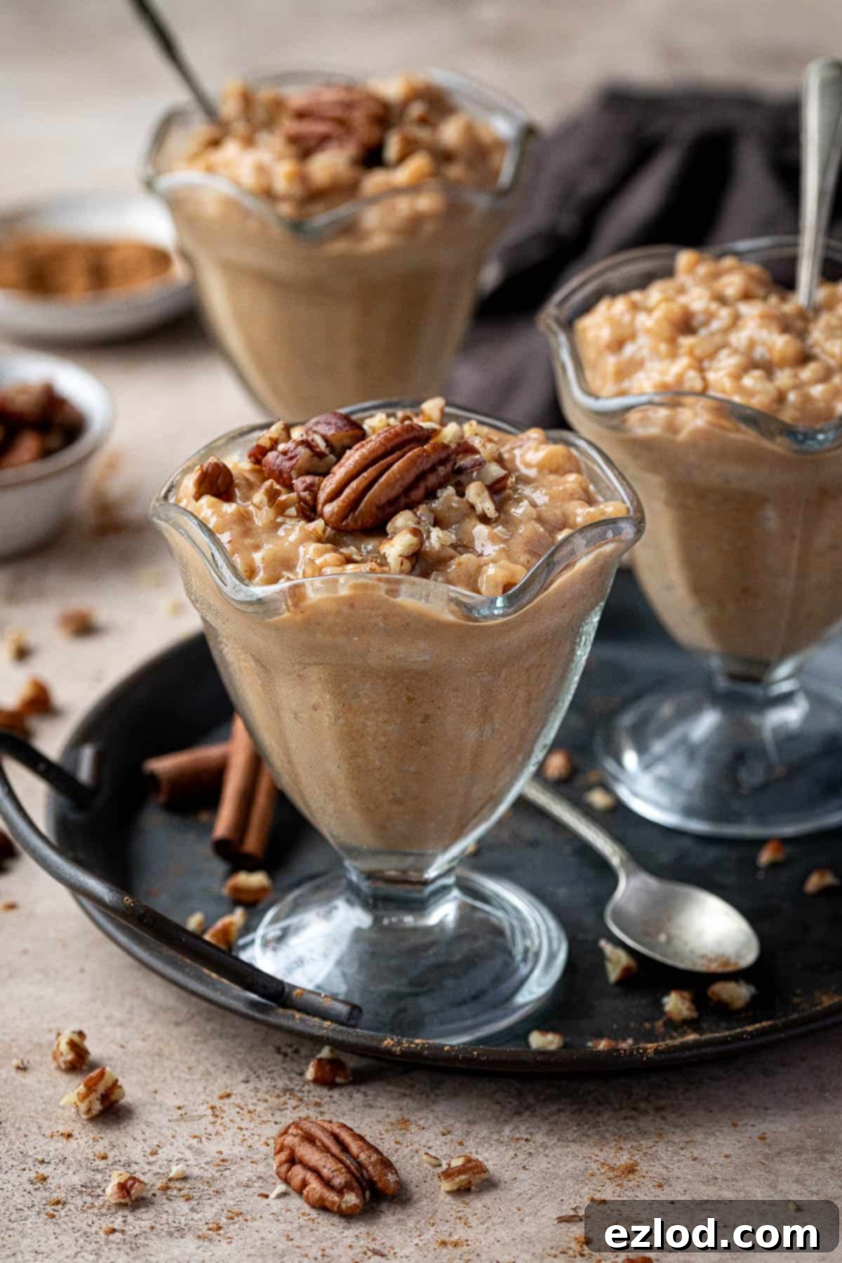 Three dessert glasses of vegan pumpkin rice pudding on a metal tray with pecans and cinnamon sticks.