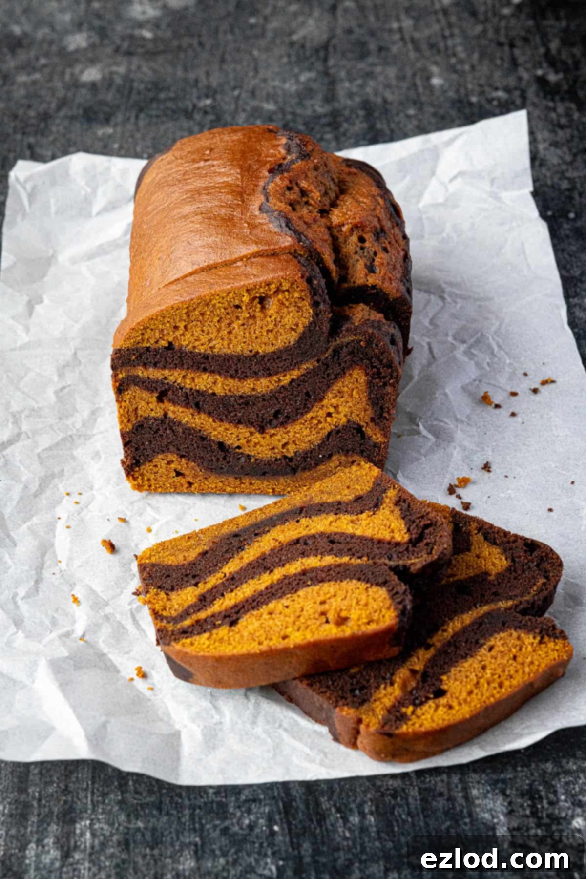 Deliciously sliced vegan pumpkin chocolate marble bread resting on baking parchment, showing off its moist crumb and intricate swirl pattern on a dark surface.