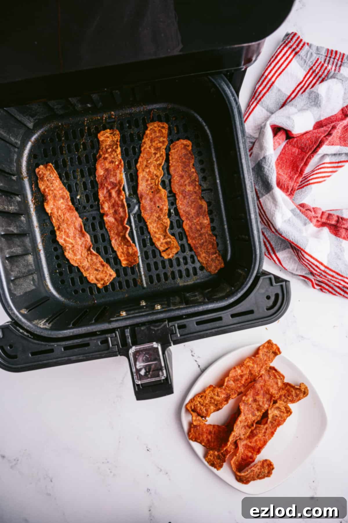Crispy Air Fryer Turkey Bacon 2 Four strips of turkey bacon cooked to crispy perfection in an air fryer basket with four more pieces sitting on a white plate nearby.