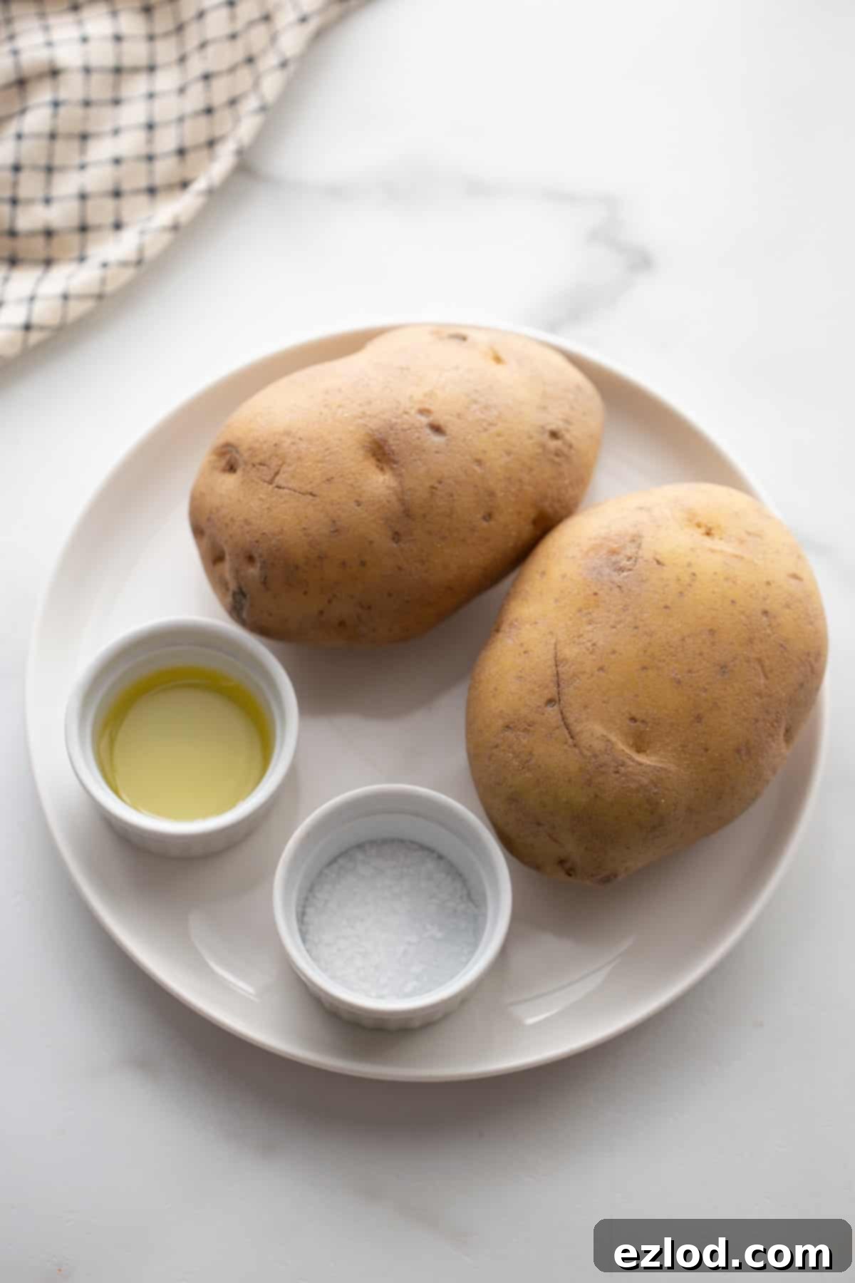 Two large raw potatoes, a bottle of avocado oil, and a small dish of salt — the simple yet crucial ingredients for making perfect homemade air fryer potato chips.