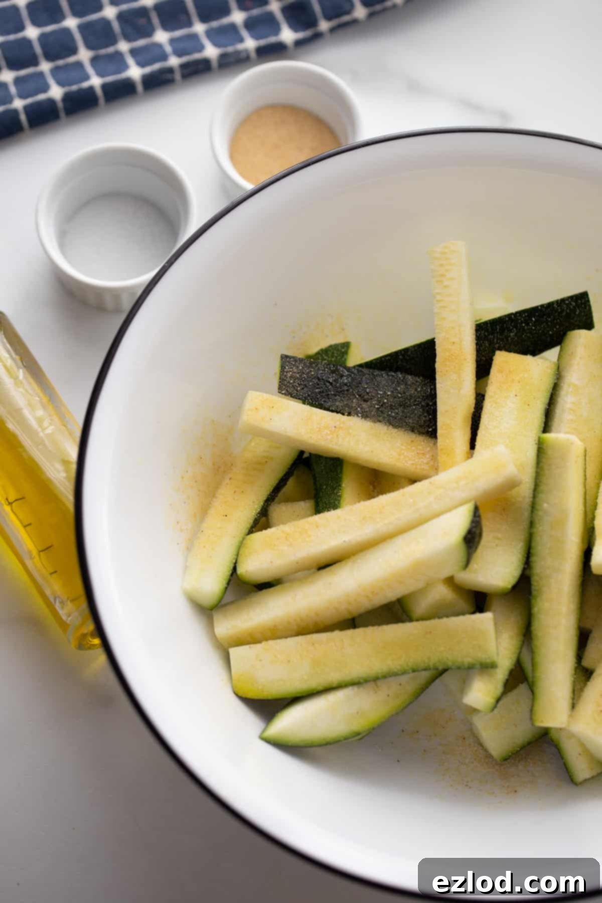A shallow white bowl with zucchini cut into sticks with salt and garlic powder sitting to the side