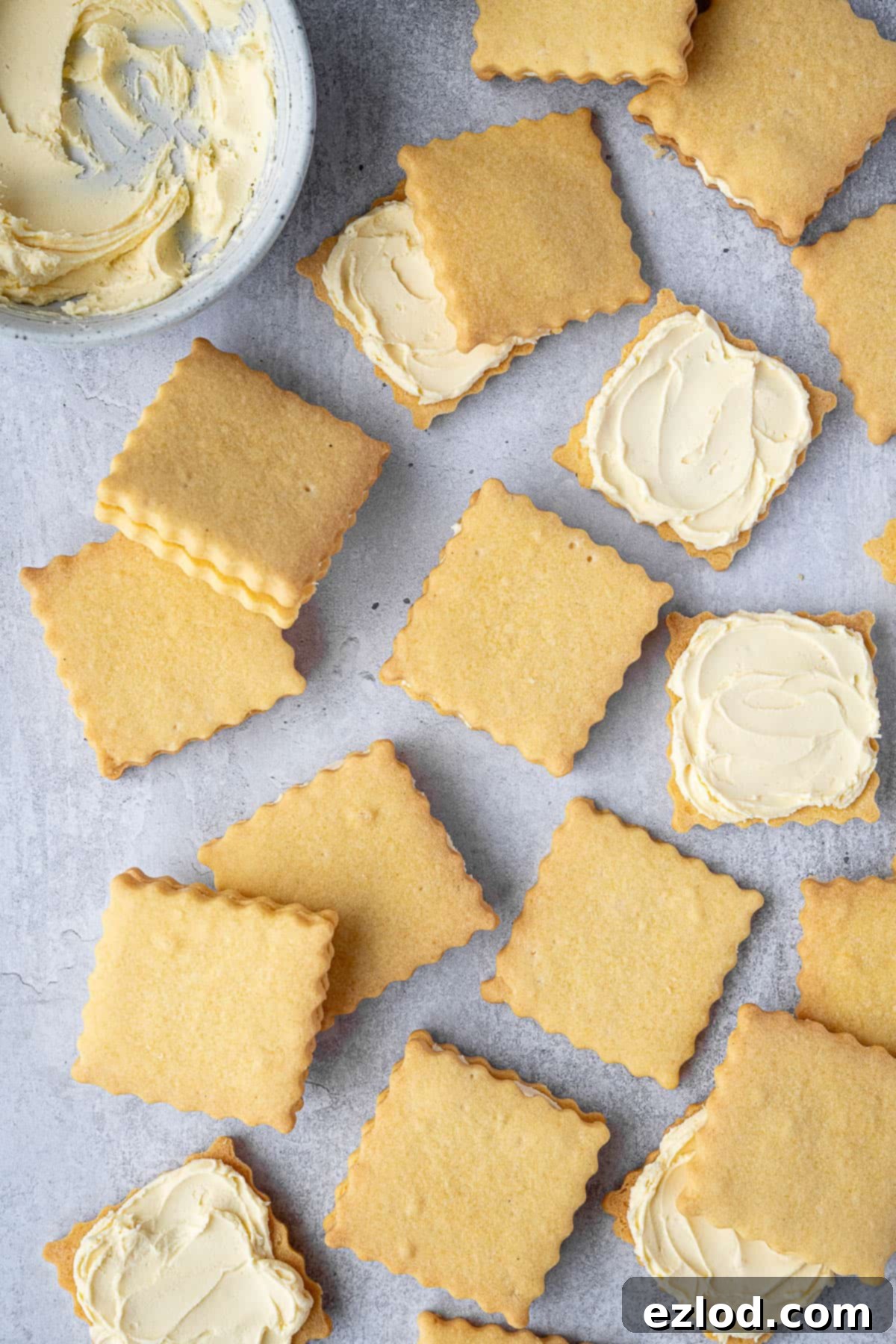 Two vegan custard cream biscuits on a white plate, with a bowl of creamy vanilla buttercream and a spatula, ready for assembly.
